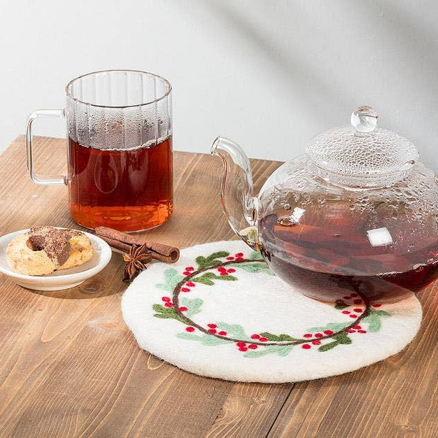 A clear glass teapot filled with tea sits on a round white doily with a red berry and green leaf border, placed on a wooden table. A matching clear glass mug with amber tea, a small dish with cookies, and a cinnamon stick are arranged nearby.