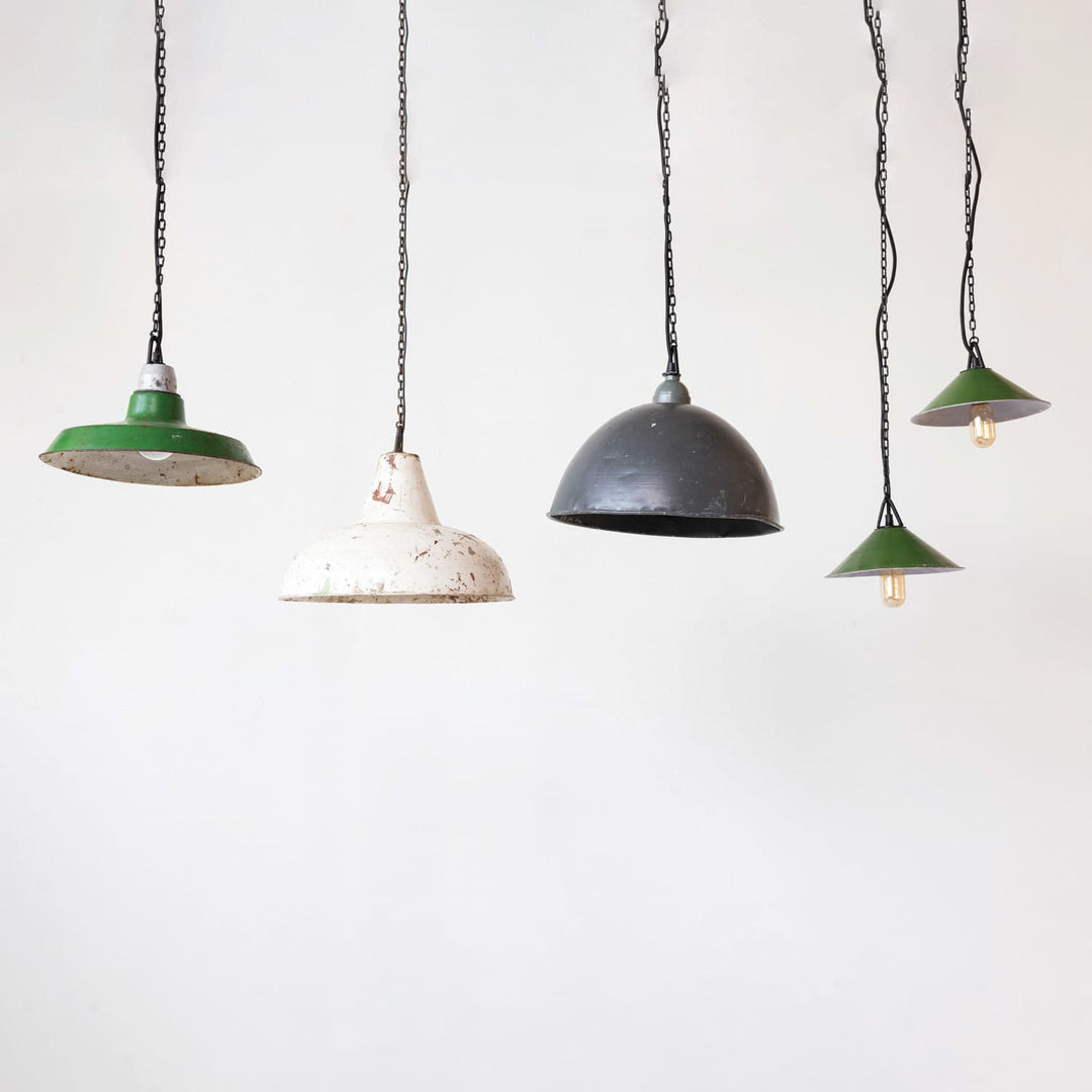Four vintage-style industrial pendant lights hanging from black chains against a plain white wall. The lights feature dome-shaped metal shades in green, distressed white, and dark gray/black, with two showing visible warm Edison bulbs.