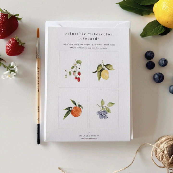 Pack of paintable watercolor notecards with fruits and flowers on a white surface with lemons, strawberries, and a pencil.