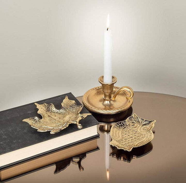 A styled tabletop scene with a lit white candle in a gold-toned cup-shaped holder, a black book, and two decorative gold-toned leaf-shaped objects on a reflective dark surface against a light neutral background.