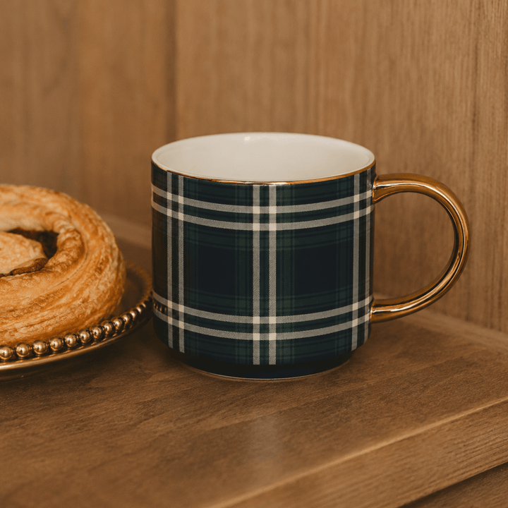 A ceramic mug with a navy blue and white plaid pattern and a gold handle, sitting on a wooden table next to a pastry.