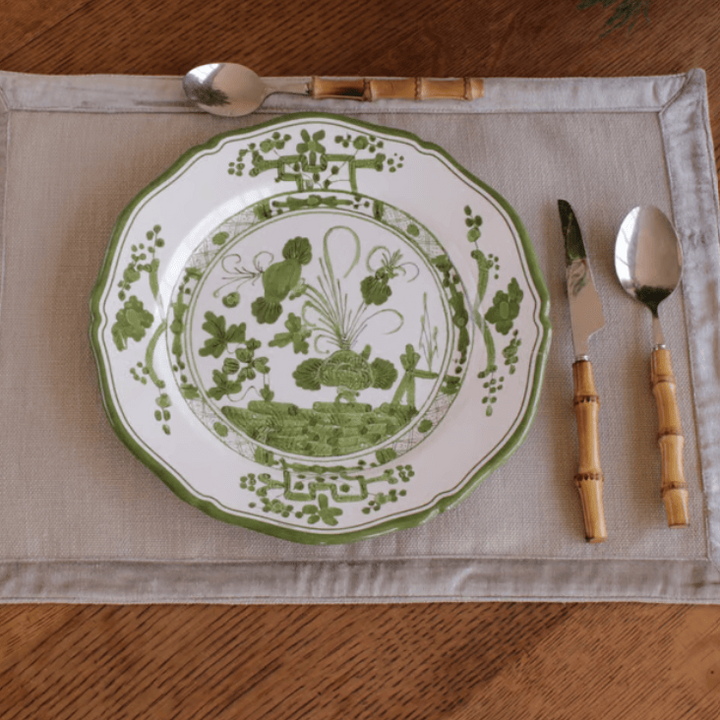 A green and white patterned melamine plate is set on a beige placemat with a bamboo-handled spoon, knife, and spoon on a wooden table.