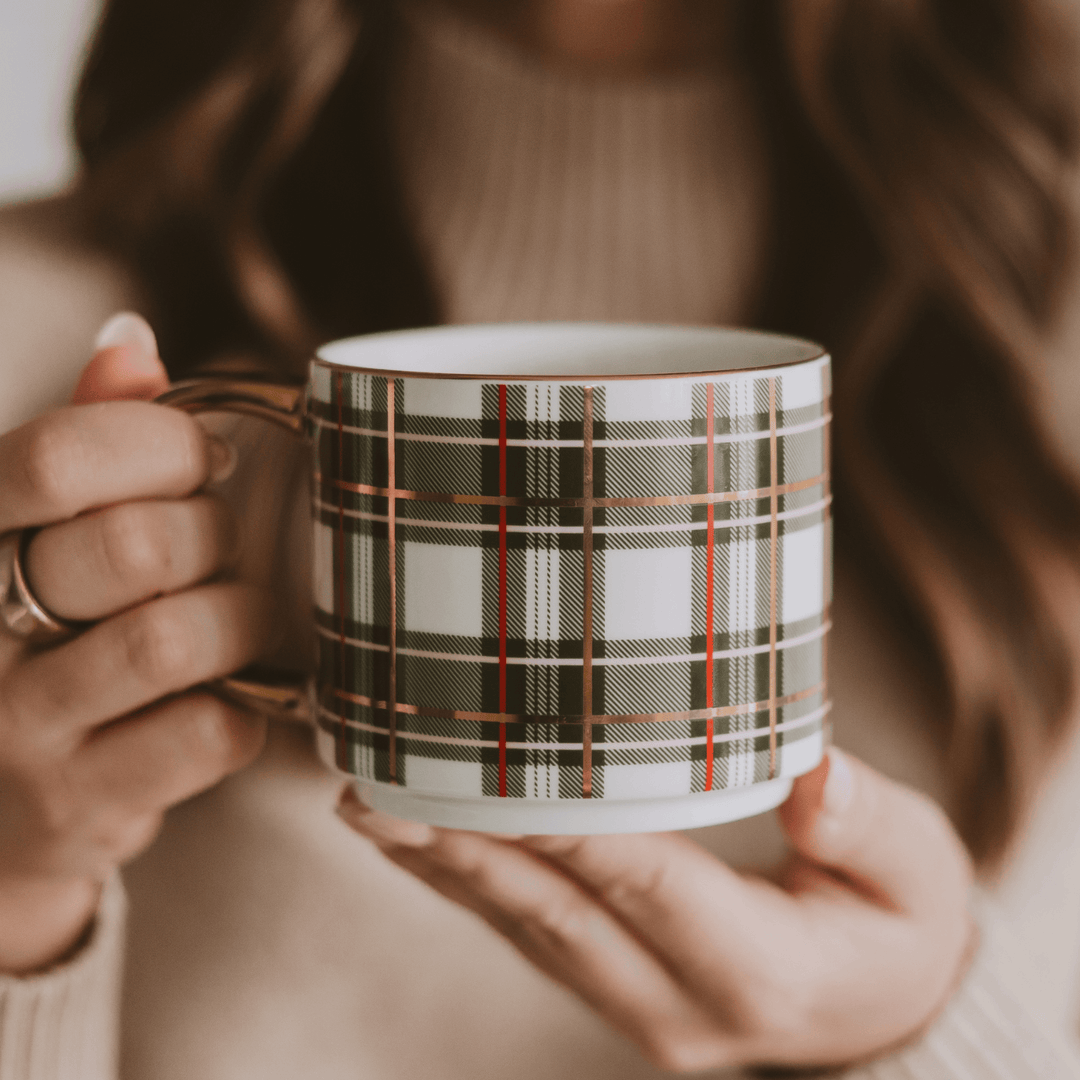 A person holding a white ceramic mug with a green, black, and gold plaid pattern, wearing a beige ribbed sweater.