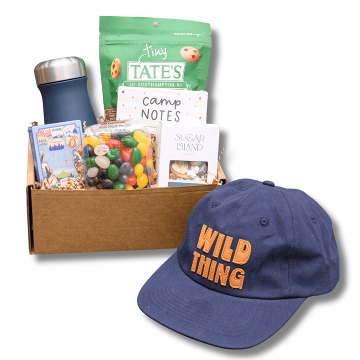 A curated gift box set featuring a navy blue baseball cap with orange 'WILD THING' embroidery, a brown cardboard box filled with snacks, a green bag of Tiny Tate's Chocolate Truffles, a notebook, and other small gift items, all on a white background.