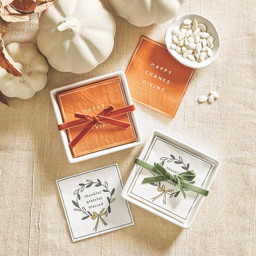 A styled flat-lay of Thanksgiving-themed cocktail napkins on a beige linen surface with white pumpkins and a small bowl of white beans. The set includes orange cocktail napkins with gold foil Happy Thanksgiving lettering tied with a red bow for gifting. 