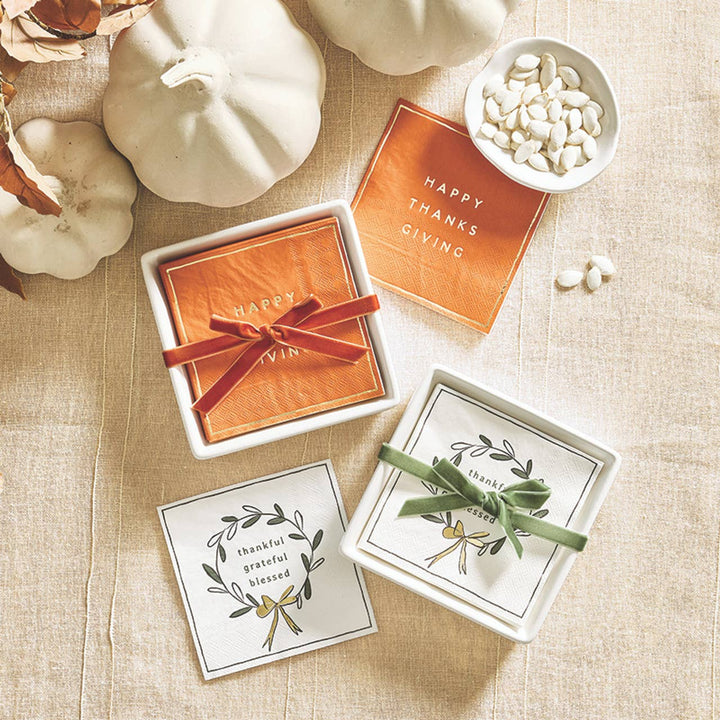 A styled flat-lay of Thanksgiving-themed cocktail napkins on a beige linen surface with white pumpkins and a small bowl of white beans. The set includes orange cocktail napkins with gold foil Happy Thanksgiving lettering tied with a red bow for gifting. 
