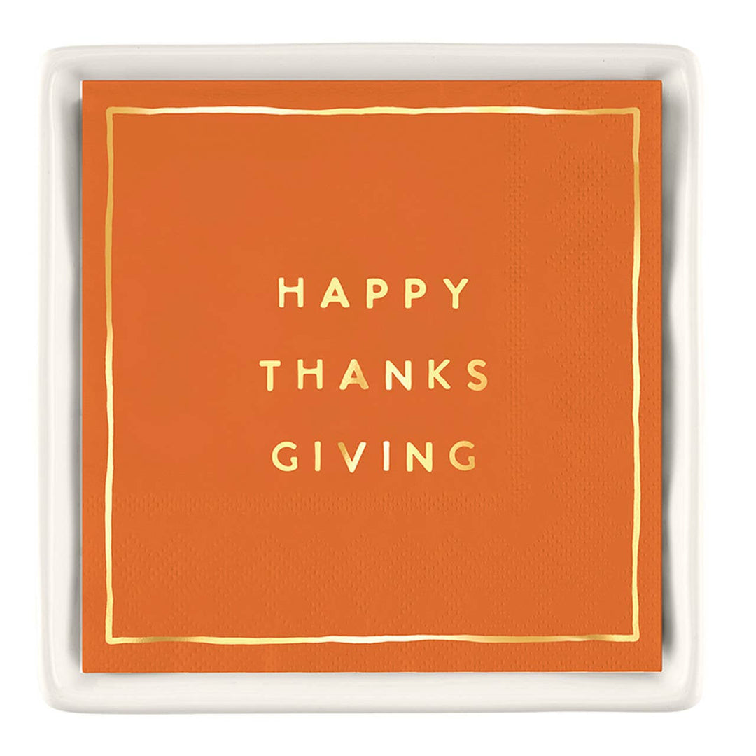 An orange square paper napkin with gold foil text reading 'HAPPY THANKS GIVING' and a thin gold border, presented on a white ceramic tray against a white background.