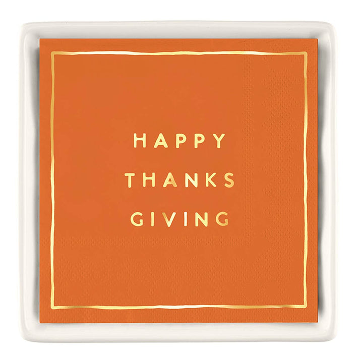 An orange square paper napkin with gold foil text reading 'HAPPY THANKS GIVING' and a thin gold border, presented on a white ceramic tray against a white background.