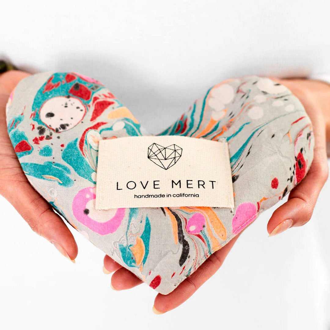 Heart-shaped eye pillow with colorful design held by hands, branded 'LOVE MERT'.