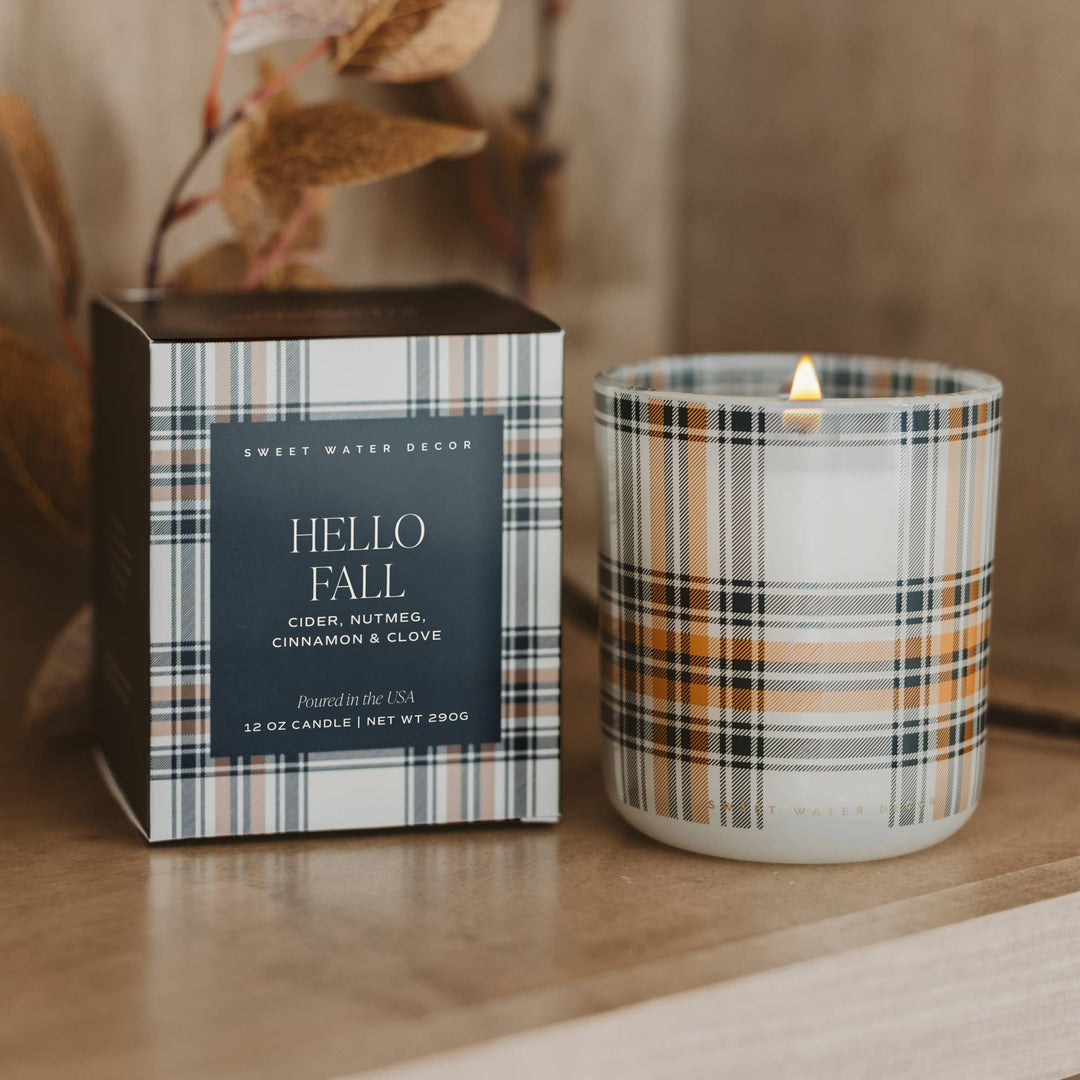 A lit scented candle in a clear glass jar with a plaid pattern sits beside its matching box on a wooden surface. The plaid features white, black, gray, and warm tan stripes, and the box has a dark charcoal label with white text reading 'HELLO FALL' and 'Sweet Water Decor'.