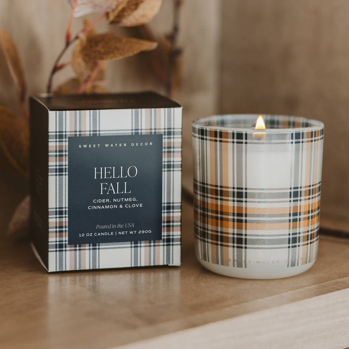 A lit scented candle in a clear glass jar with a plaid pattern sits beside its matching box on a wooden surface. The plaid features white, black, gray, and warm tan stripes, and the box has a dark charcoal label with white text reading 'HELLO FALL' and 'Sweet Water Decor'.