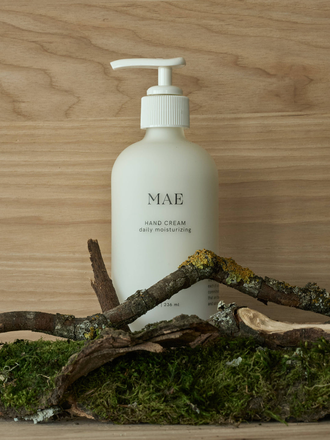 Hand cream bottle labeled 'MAE' on a wooden surface with branches and moss.