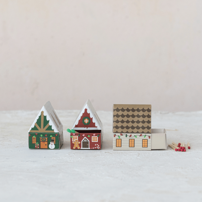 Three small decorative Christmas-themed matchboxes lined up on a light surface against a pale background. The matchboxes are shaped like gingerbread houses, with one green and one red decorated house and one beige house with a brown patterned roof.