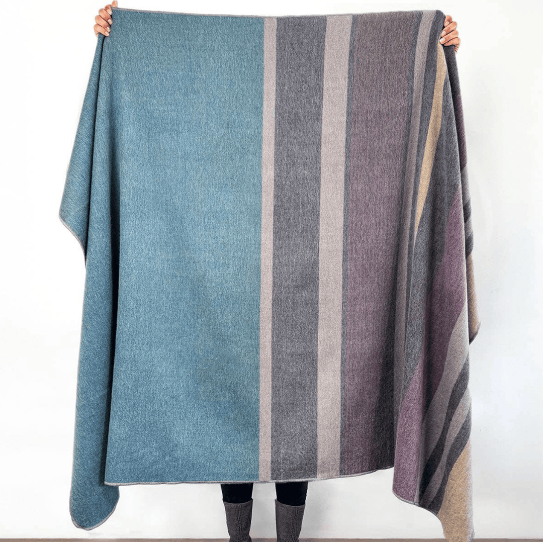 A large striped wool blanket or throw held up by a person against a plain white wall. The blanket features vertical stripes in muted blue, gray, taupe, and dusty purple, with the person's hands and boots visible holding the corners.