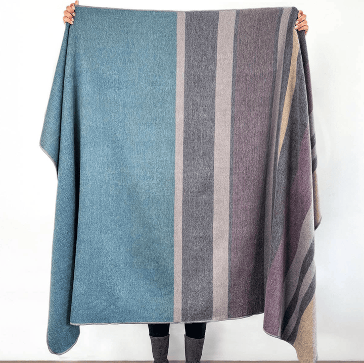 A large striped wool blanket or throw held up by a person against a plain white wall. The blanket features vertical stripes in muted blue, gray, taupe, and dusty purple, with the person's hands and boots visible holding the corners.