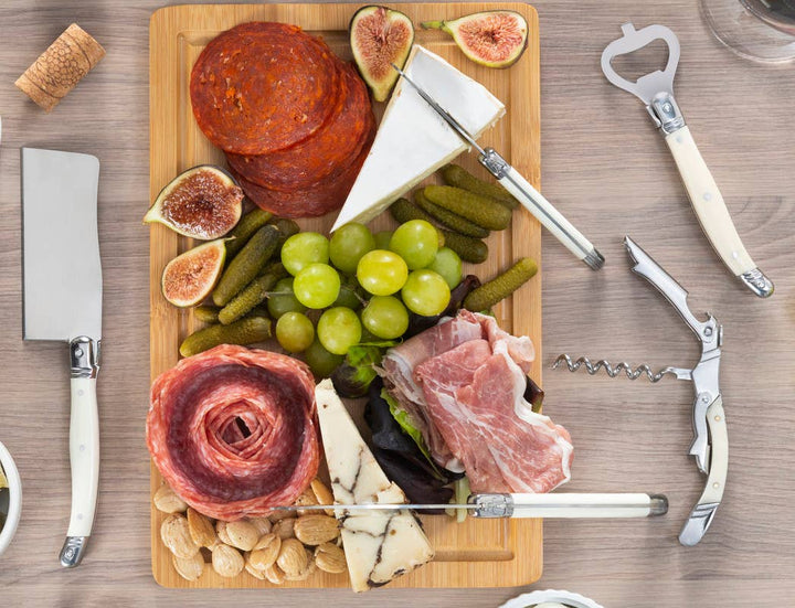 Wooden cutting board with assorted meats, cheeses, and fruits on a wooden table with faux ivory handled bottle opener and corkscrew