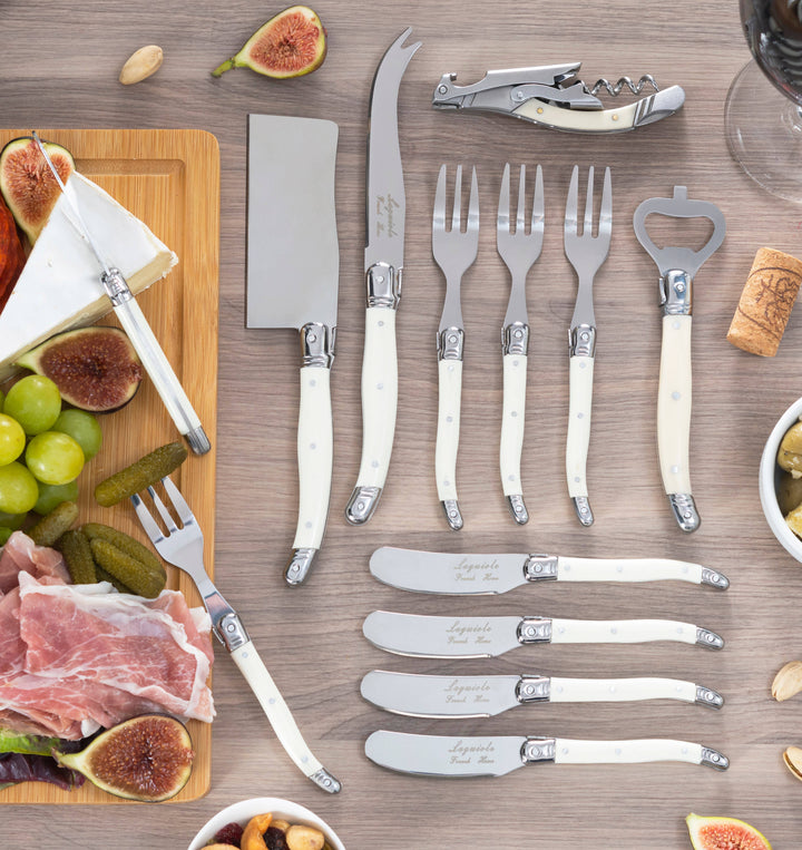 Set of knives, forks, and cheese knives with faux ivory handles on a wooden surface with food items.