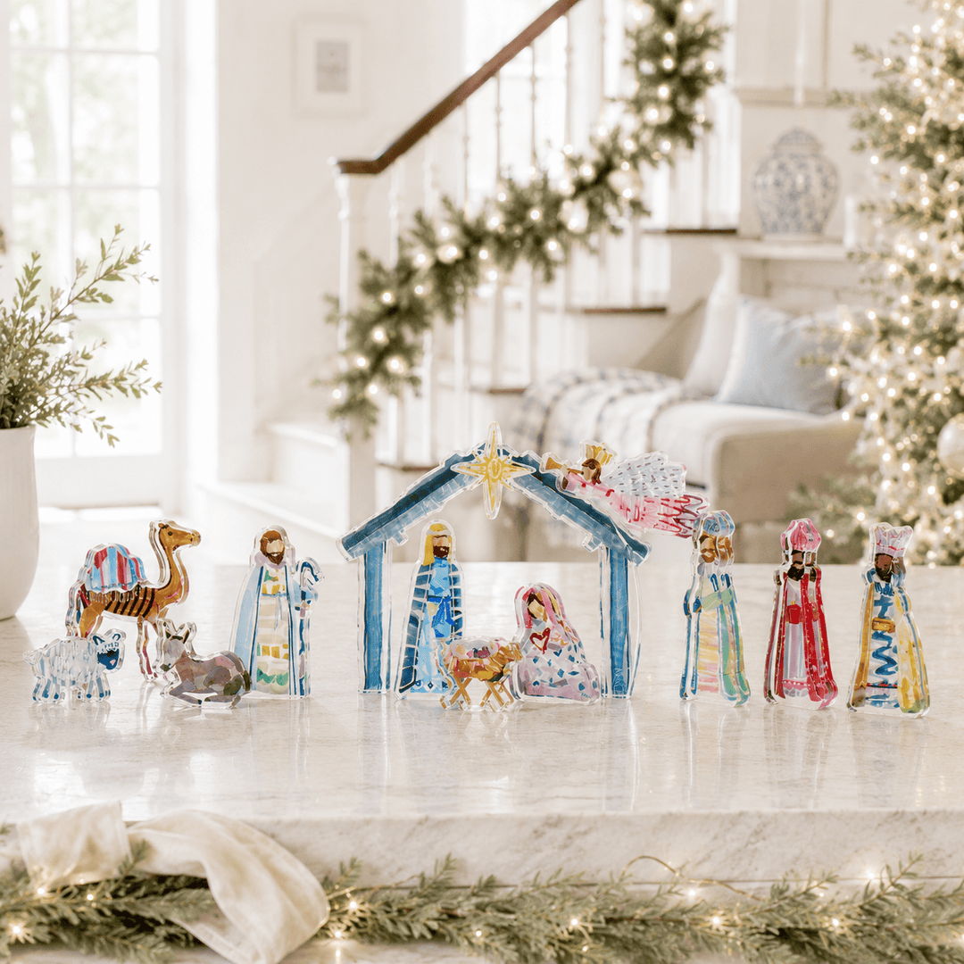 A colorful Christmas nativity scene tabletop decoration set displayed on a marble countertop in a bright, decorated living room. The set includes a blue stable with a gold star, Mary and Joseph in blue robes, baby Jesus, three wise men in colorful robes, a camel, a donkey, and a sheep.