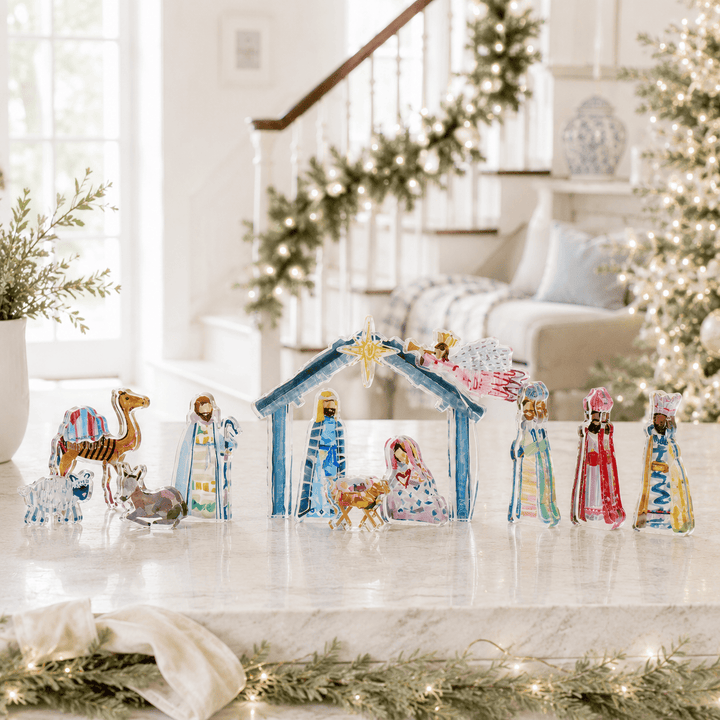 A colorful Christmas nativity scene tabletop decoration set displayed on a marble countertop in a bright, decorated living room. The set includes a blue stable with a gold star, Mary and Joseph in blue robes, baby Jesus, three wise men in colorful robes, a camel, a donkey, and a sheep.