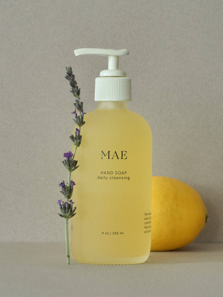Yellow bottle of MAE hand soap with a pump, lavender sprig, and lemon on a neutral background