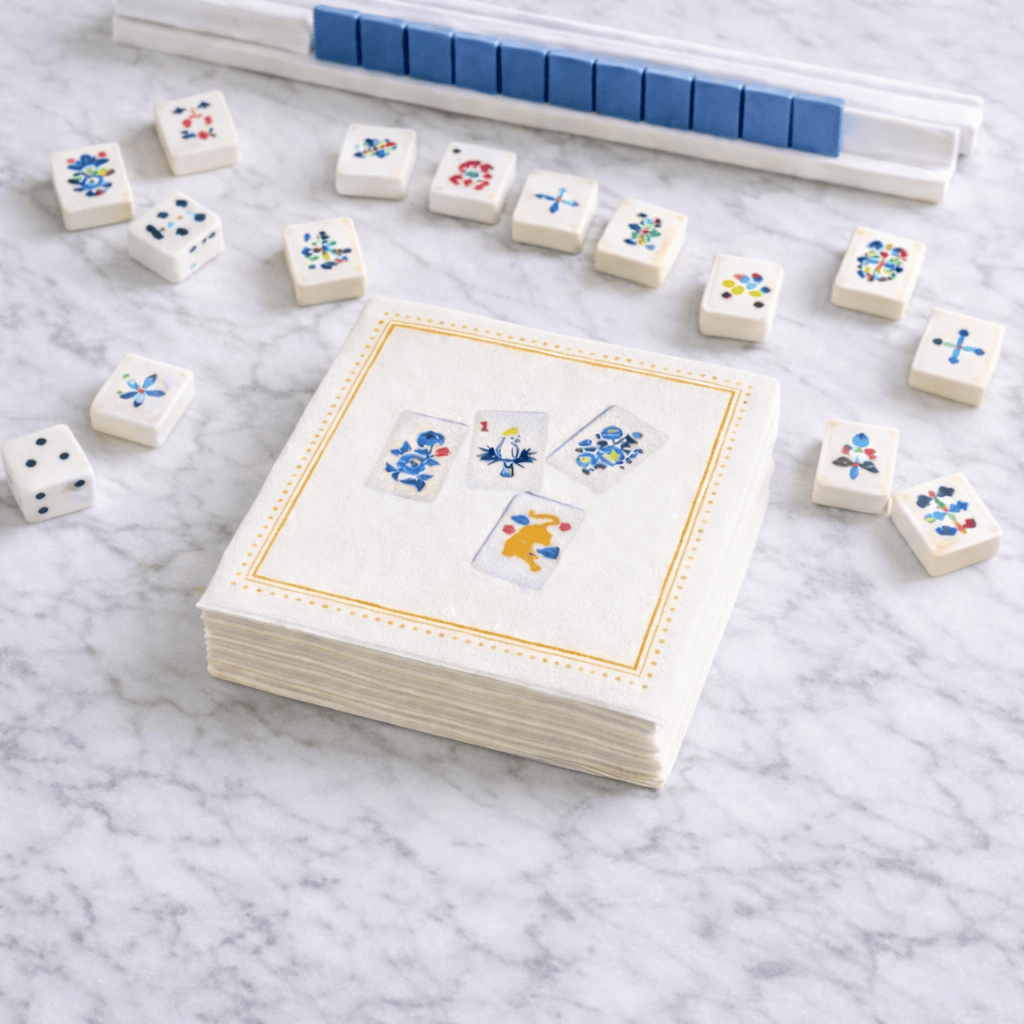A stack of white paper napkins with a gold border and printed mahjong designs sits on a white marble surface, surrounded by small white dice and a blue-and-white game board in the background.