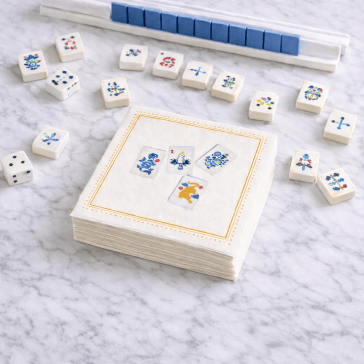 A stack of white paper napkins with a gold border and printed mahjong designs sits on a white marble surface, surrounded by small white dice and a blue-and-white game board in the background.