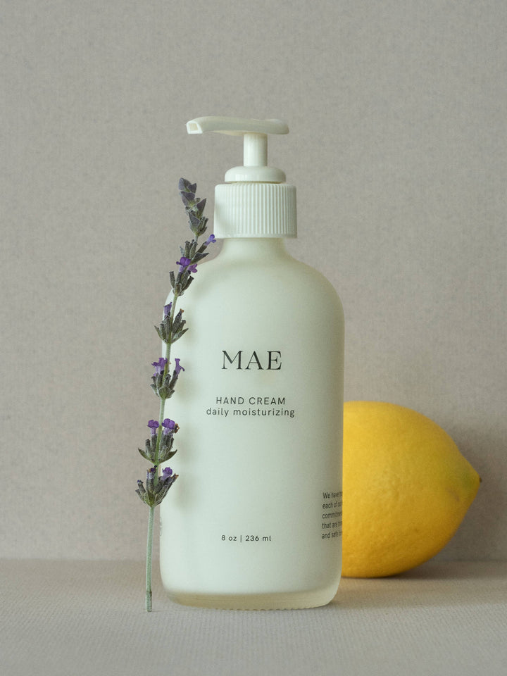 Hand cream bottle labeled 'MAE' with a lemon and lavender sprig on a neutral background