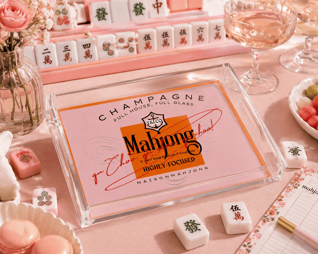A clear acrylic Mahjong tray with a pink and orange printed insert reading 'CHAMPAGNE FULL HOUSE FULL GLASS' and 'Maison Mahjong', styled on a pink tabletop with mahjong tiles, a glass of rosé wine, flowers, and small pink accessories.