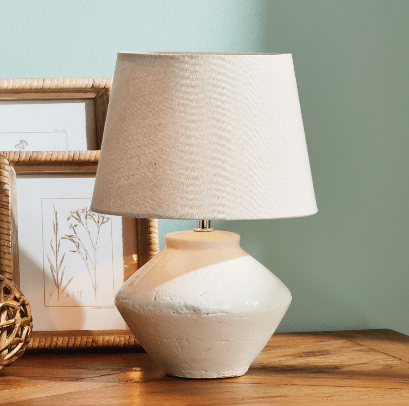 Table lamp with a beige shade on a wooden surface against a light blue wall.