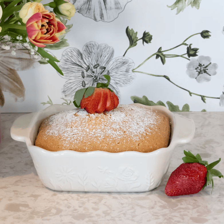 A baked loaf of bread or pastry in a white ceramic baking dish, topped with powdered sugar and a sliced strawberry, set on a patterned surface with a floral backdrop and a whole strawberry beside the dish.