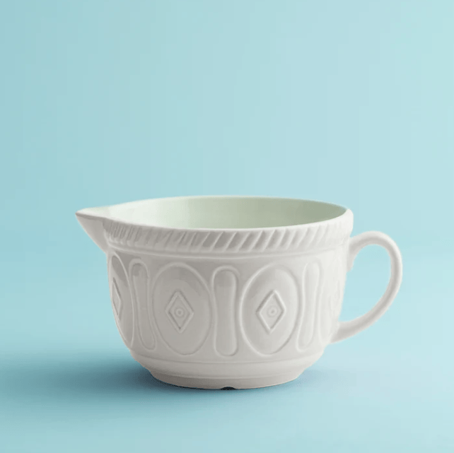 A white ceramic batter bowl with embossed geometric patterns and a small handle, set against a solid light blue background.