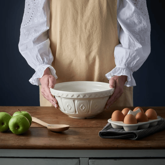 A person in a white blouse and beige apron holds a large cream-colored ceramic mixing bowl with embossed geometric patterns. The bowl is set on a rustic wooden table with green apples, a wooden spoon, and a carton of brown eggs against a dark navy background.