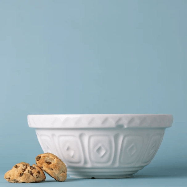 A white ceramic mixing bowl with a textured geometric pattern, shown on a light blue background with two chocolate chip cookies in the foreground.