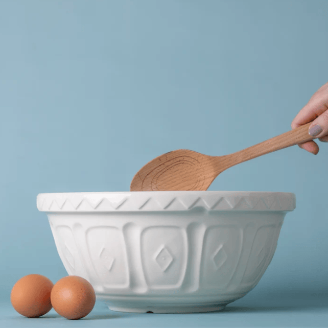 A white ceramic mixing bowl with embossed diamond patterns sits on a light blue surface, with two brown eggs beside it and a wooden spoon being held over the bowl by a visible hand.