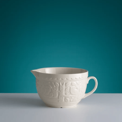 An off-white ceramic pitcher with embossed floral patterns, set against a teal background and a light gray surface.