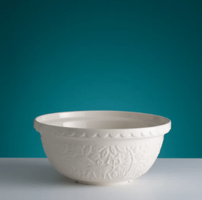 An off-white ceramic mixing bowl with embossed floral patterns and a scalloped rim, set against a teal background on a light gray surface.