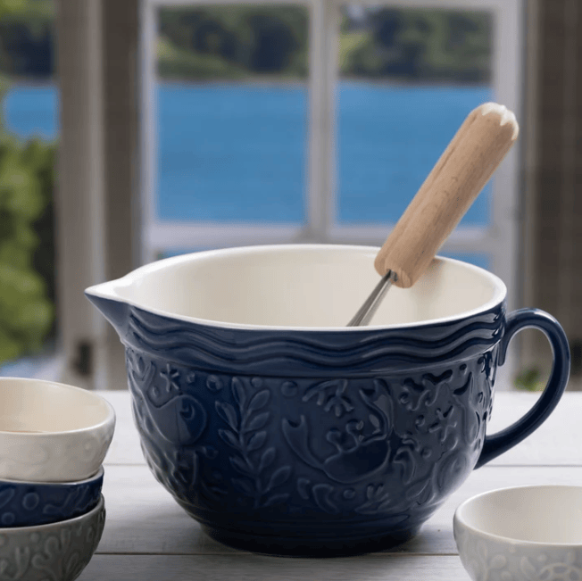 A large navy blue ceramic batter bowl with an embossed floral pattern, a white interior, a pouring spout, and a wooden-handled metal whisk resting inside. The bowl sits on a light tabletop with a blurred window and blue water in the background, alongside smaller matching bowls.
