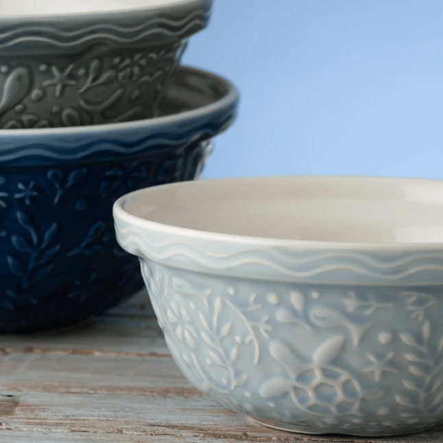 A close-up of a light blue-gray ceramic bowl with embossed floral patterns and a wavy rim, with two darker bowls stacked in the background on a rustic wooden surface against a soft blue backdrop.