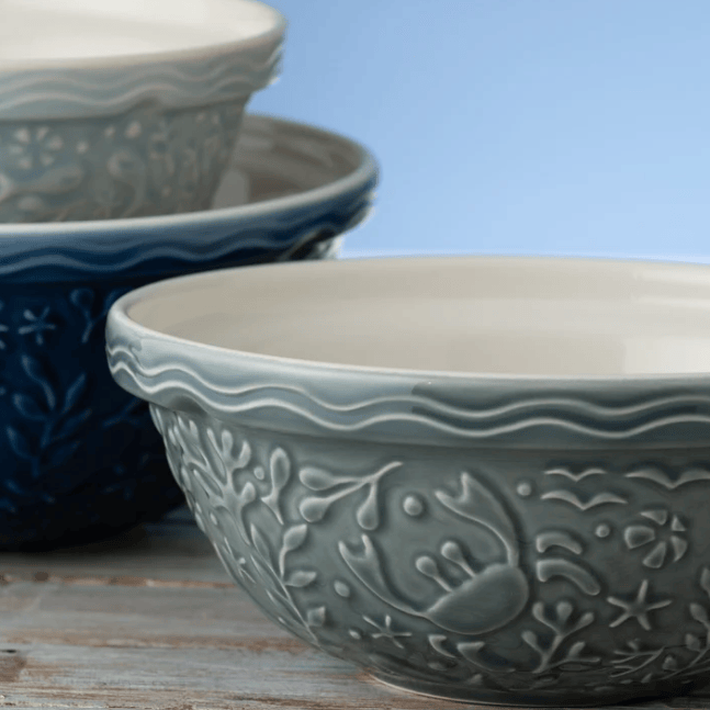 A close-up of a ceramic bowl with embossed floral and leaf patterns, shown in the foreground with two additional bowls stacked behind it on a rustic wooden surface against a soft blue background.
