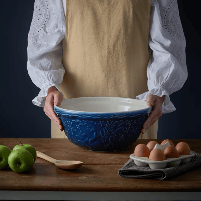 A person wearing a white blouse and tan apron holds a large ceramic mixing bowl with a deep blue embossed floral border and a white interior. The bowl sits on a wooden table with green apples, a wooden spoon, and a carton of brown eggs nearby against a dark background.