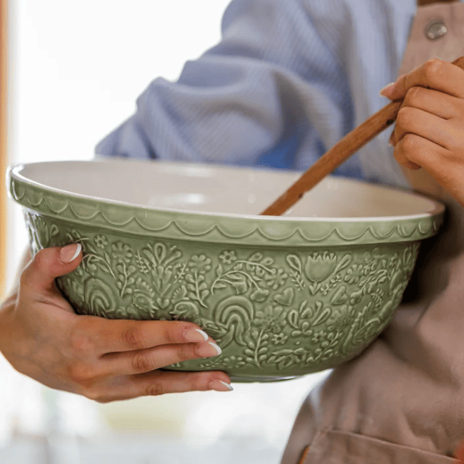 A person holding a large green ceramic mixing bowl with embossed floral detailing, mixing ingredients with a wooden spoon.
