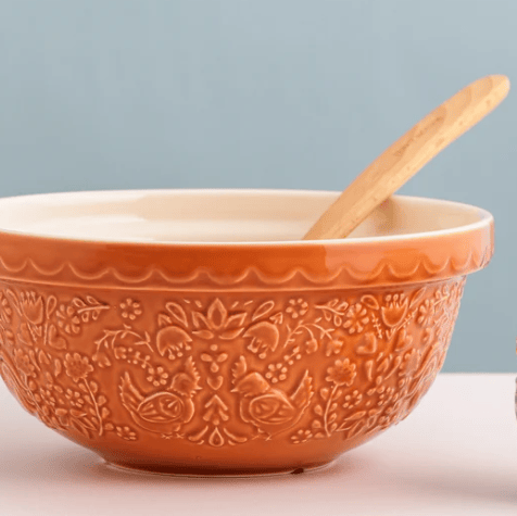 An orange ceramic mixing bowl with embossed floral and bird patterns, featuring a scalloped rim and a light wood spoon inside, set against a soft blue-gray background.