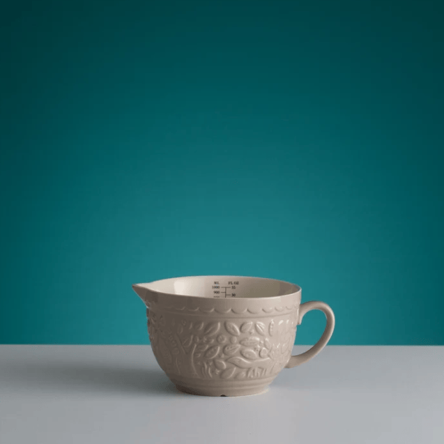 An off-white ceramic pitcher with embossed floral patterns and a handle, set against a solid teal background and a light gray surface.