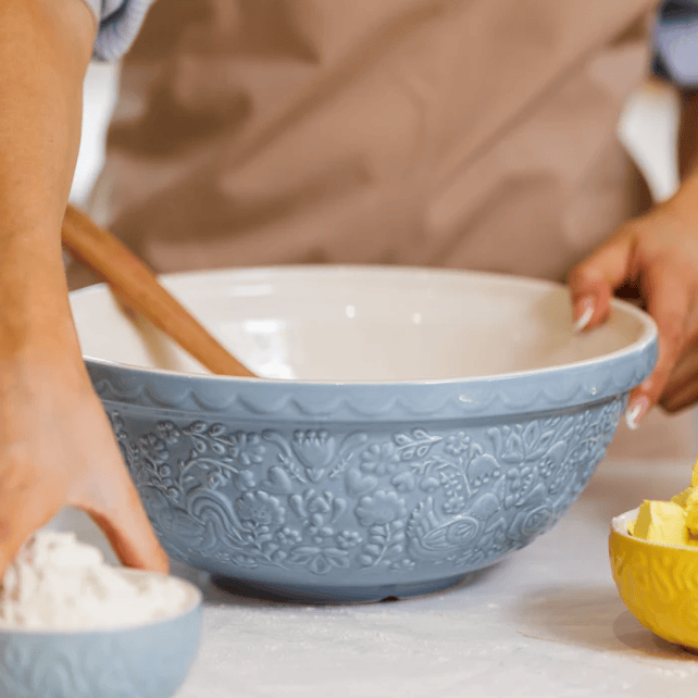 A large light blue ceramic mixing bowl with embossed floral detailing and a white interior, shown on a countertop with hands reaching in and baking ingredients nearby.
