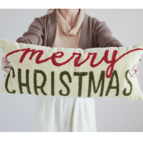 A person holding a long rectangular decorative pillow with the words 'merry CHRISTMAS' in red and dark green on a cream background.