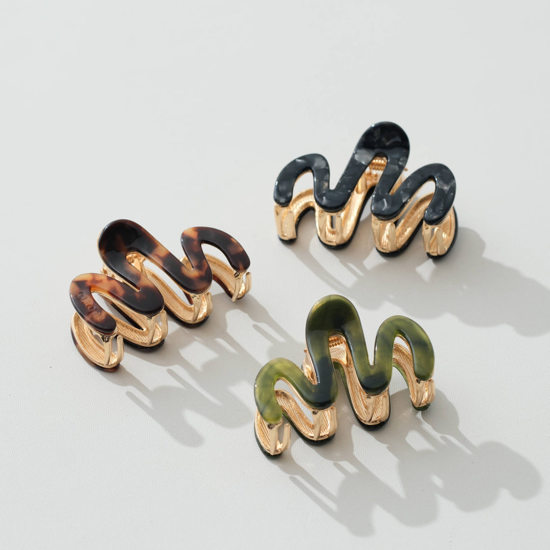 Three wavy spiral hair claw clips with gold-tone metal and tortoiseshell, black, and olive green enamel, arranged on a light gray background.