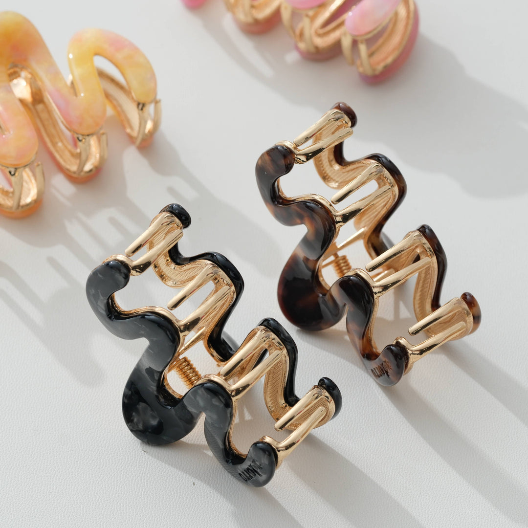 Three wavy claw hair clips with gold-tone metal and glossy black, tortoiseshell, and pastel pink resin, arranged on a light gray surface.