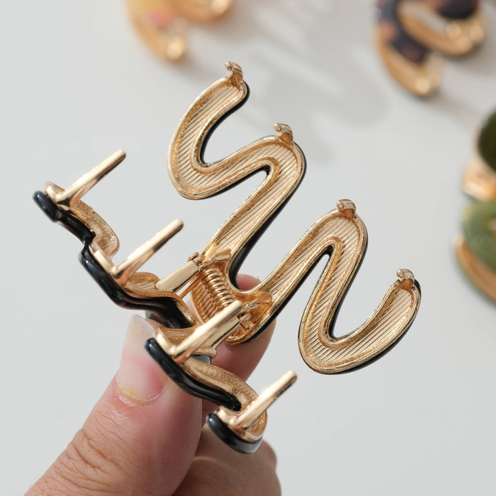 A close-up of a hand holding a gold-toned claw hair clip with black enamel accents against a light gray background.