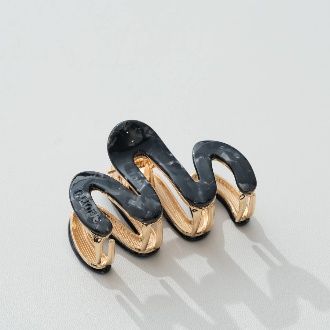 A sculptural spiral hair claw clip featuring glossy black and translucent dark gray enamel sections over a polished gold-tone metal base, photographed on a light gray surface.