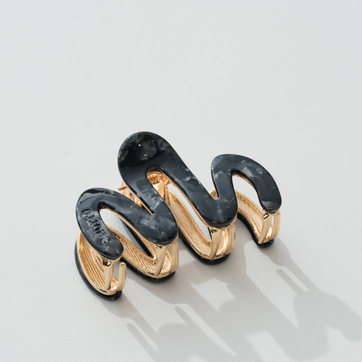 A sculptural spiral hair claw clip featuring glossy black and translucent dark gray enamel sections over a polished gold-tone metal base, photographed on a light gray surface.